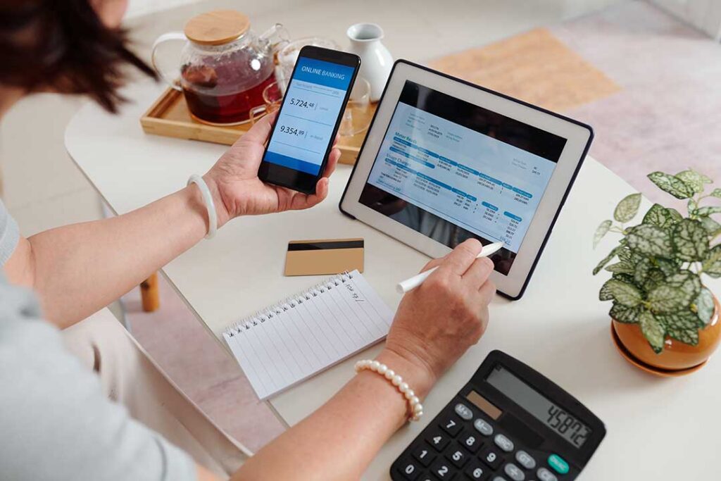 A smiling older couple reviews financial papers at their kitchen table with a laptop and coffee.